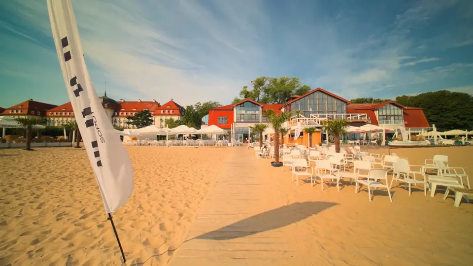 Plaża MUDO w złotym świetle: pomost na piasku, białe meble plażowe, palmy i budynek MUDO