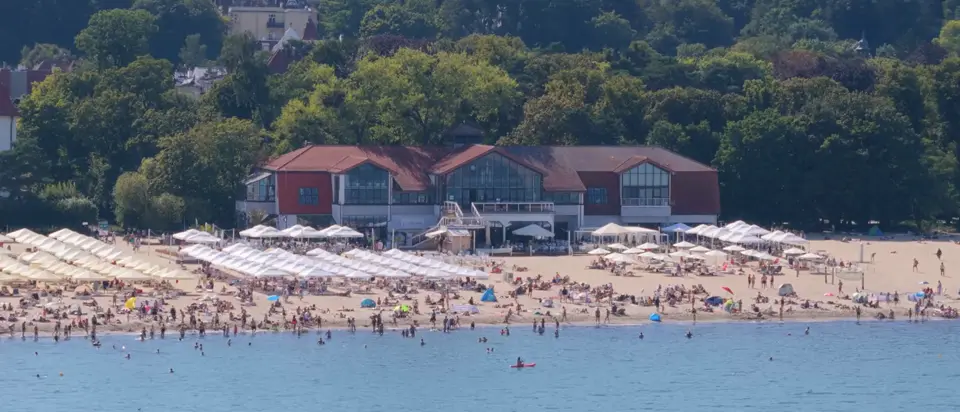 Przelot dronem wzdłuż plaży MUDO w Sopocie: budynek z czerwonym dachem, taras z parasolami i park za budynkiem