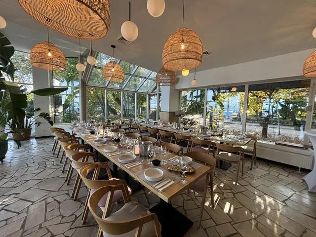 Sala restauracyjna w oranżerii: długie drewniane stoły, wiklinowe lampy i przeszklony dach z widokiem na zieleń