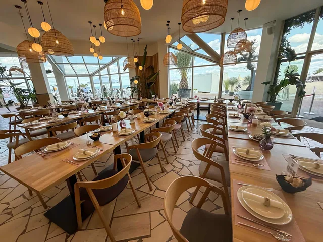 Sala restauracyjna nakryta do bankietu: rzędy jasnych drewnianych stołów pod wiklinowymi lampami, przeszklona ściana od strony plaży