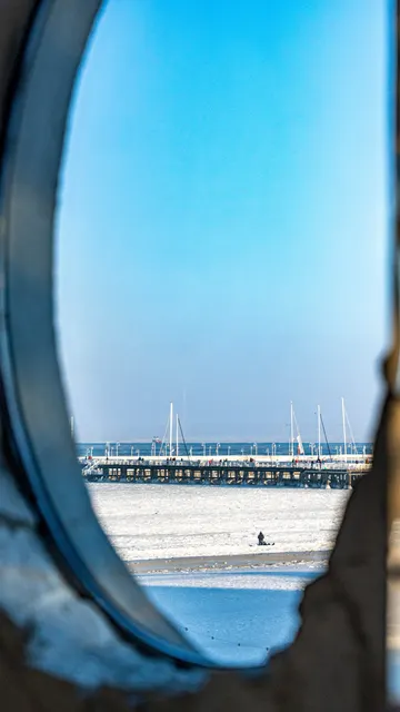 Widok przez okrągły bulaj na ośnieżoną plażę i molo w słoneczny zimowy dzień