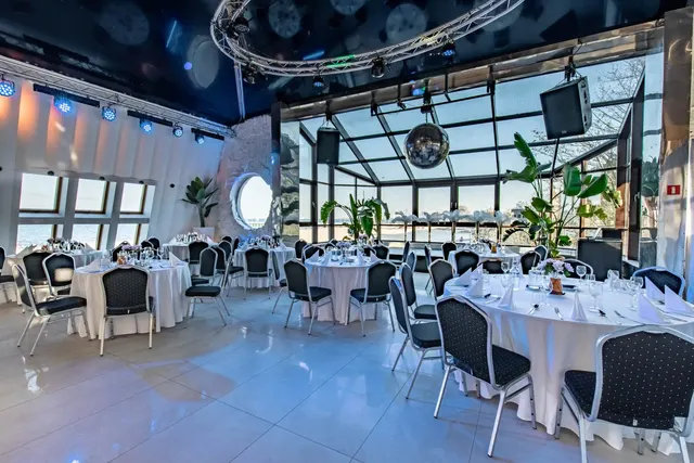 Sala klubowa nakryta do kolacji: białe okrągłe stoły, zieleń w donicach i przeszklenia od strony morza