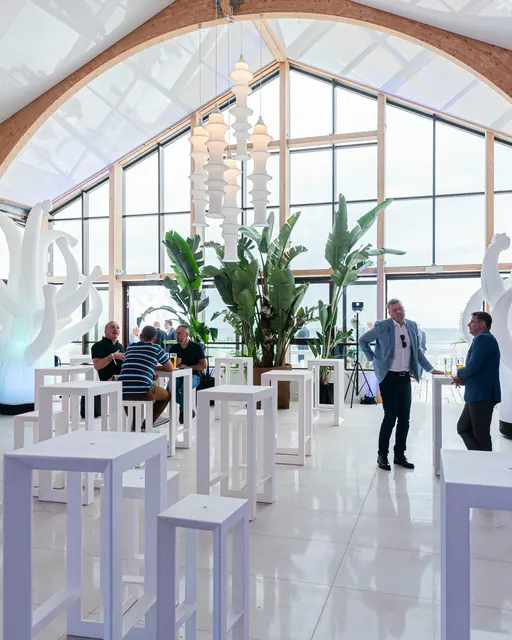 Sala restauracyjna z wysokim przeszkleniem i palmami, białe stoliki koktajlowe i goście przy wejściu