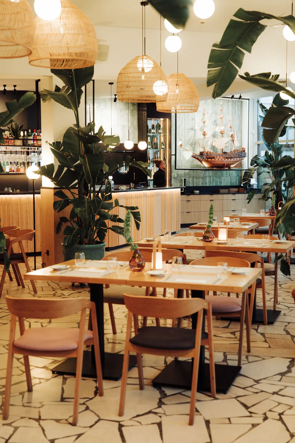 Sala restauracyjna ze stolikami, roślinami i barem w głębi, osoba przy barze