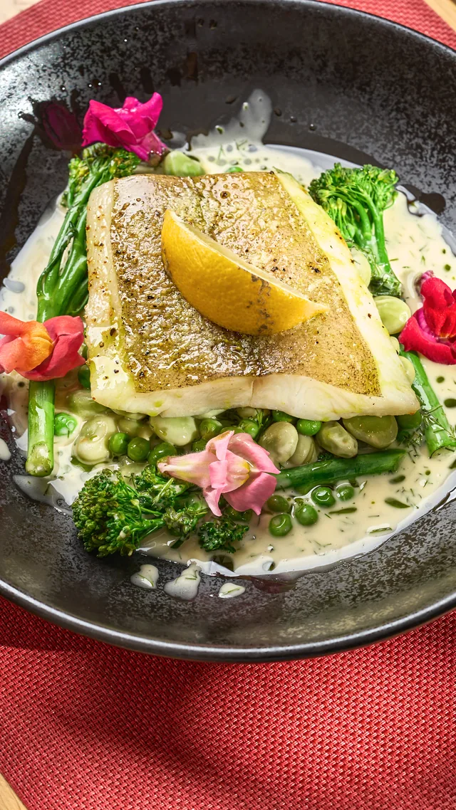 Halibut z zielonymi warzywami, sosem i grillowaną cytryną na czarnym talerzu, w tle liście palmy